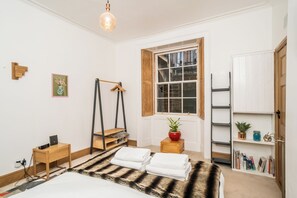 Ein Schlafzimmer mit Bett, Nachttisch, Lampe, Fenster und einem Regal mit Büchern und Pflanzen.
