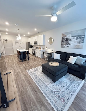 A modern living room with a dark sofa, a coffee table, and a kitchen in the background. - New Modern Luxe 1 Bed 1 Bath w/ Resort Amenities (Waukee)