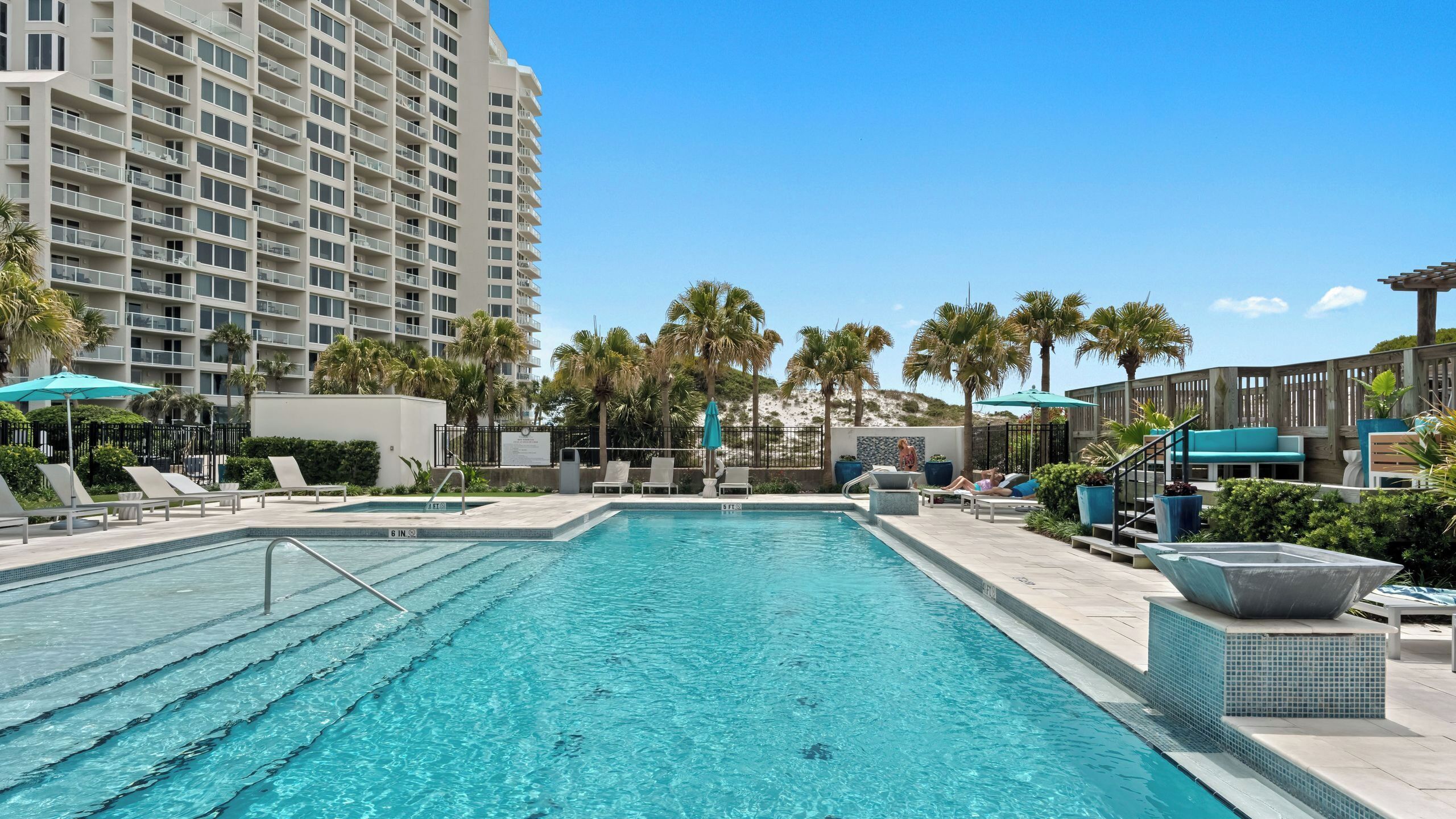 A swimming pool with lounge chairs and umbrellas, surrounded by palm trees and a high-rise building.