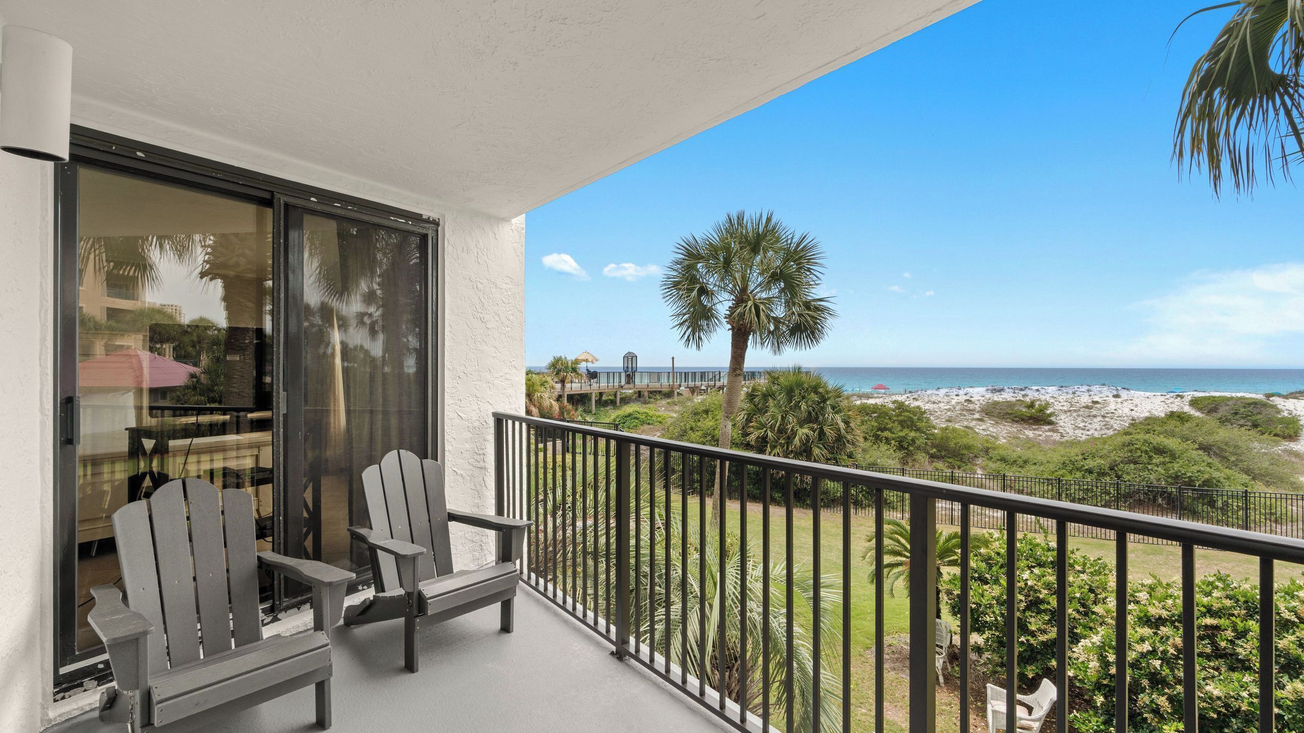 A balcony with two Adirondack chairs, a glass door, and a view of the beach and ocean.