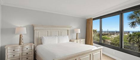 A bedroom with a large bed, two bedside tables, a view of the beach, and a window with curtains.