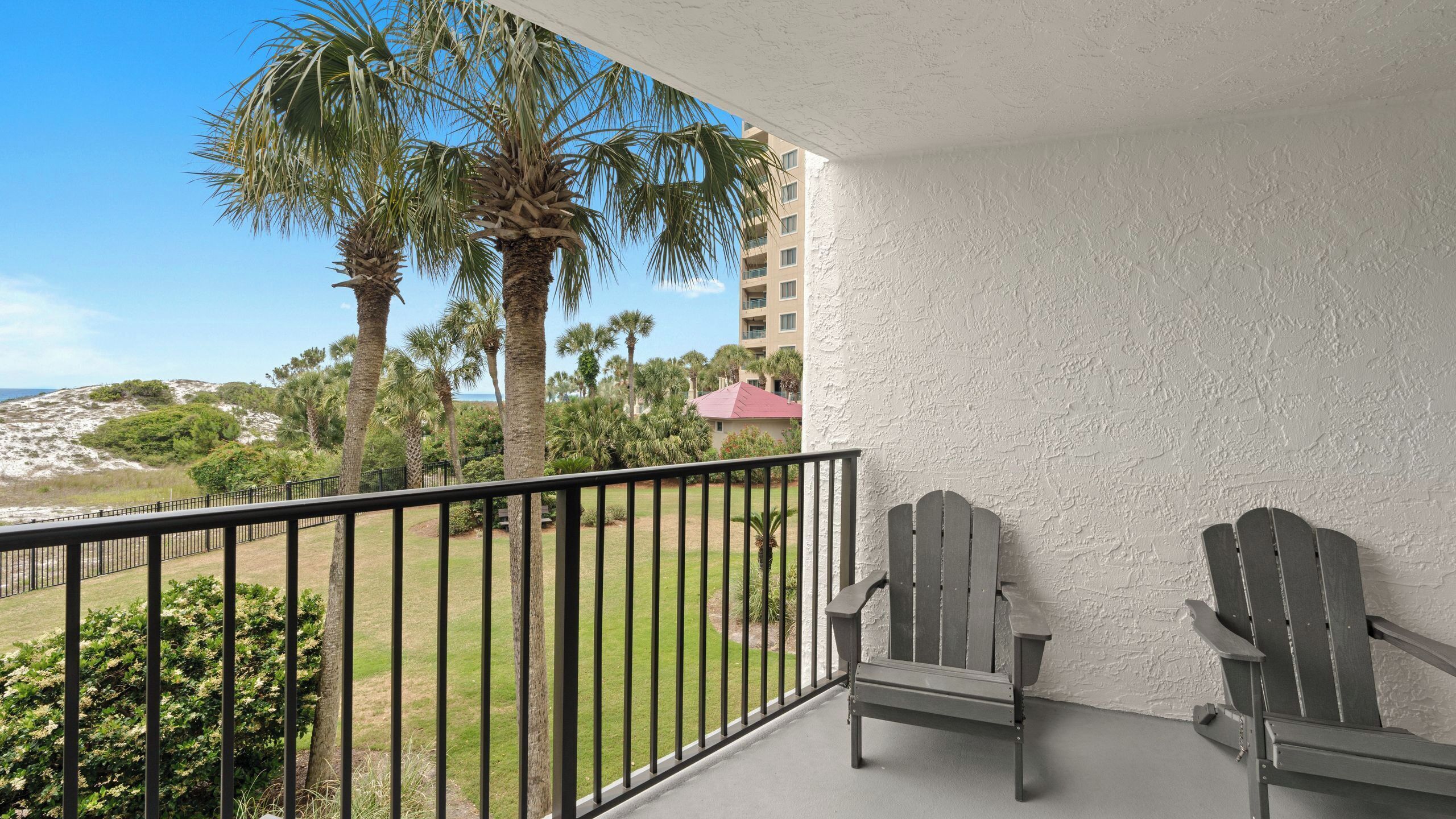A balcony with two Adirondack chairs, a view of a beach, palm trees, and a building with a red roof.