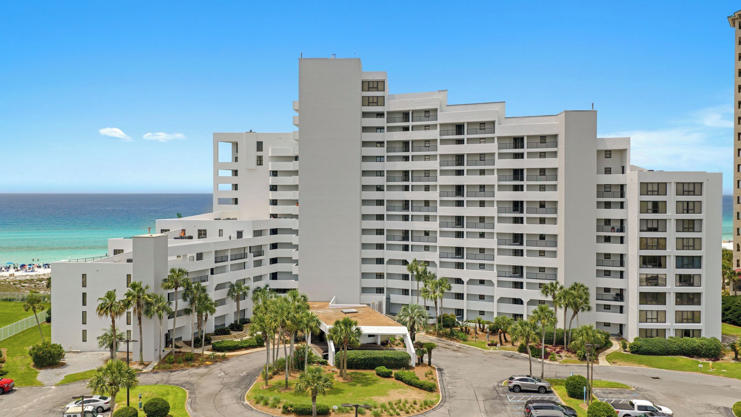 A multi-story hotel with balconies, a parking area, and a view of the ocean.