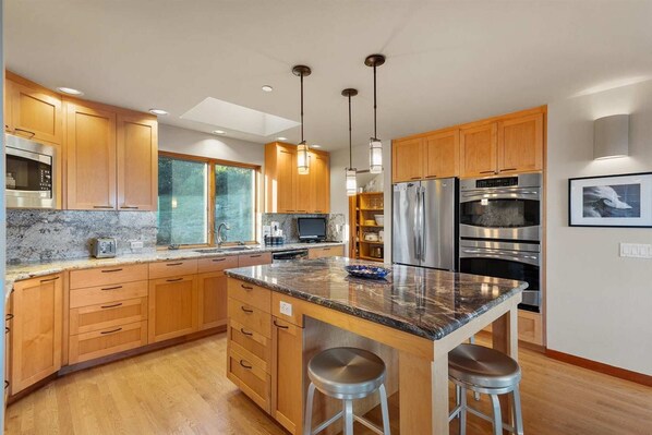 A modern kitchen with wooden cabinets, stainless steel appliances, a marble countertop, and pendant lighting.