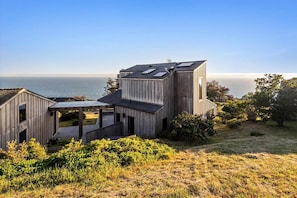 A modern wooden house with a view of the sea, featuring a large deck and a covered outdoor area.