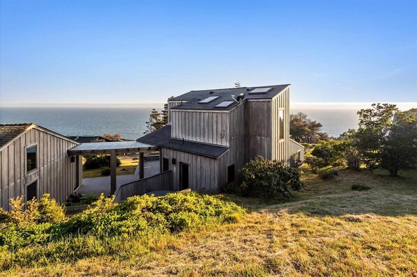 A modern wooden house with a view of the sea, featuring a large deck and a covered outdoor area.