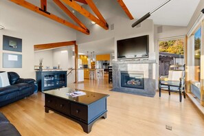 A spacious living room with a fireplace, wooden beams, a TV, and a kitchen in the background.