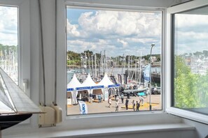 A view through a window of a marina with sailboats, tents, and a clear sky. - La Perle du Port : Vue Panoramique et Terrasse (La Trinité-sur-Mer)