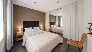 Quarto de hotel moderno com uma cama grande, mesas de cabeceira com luminárias, um espelho e uma cadeira de vime.