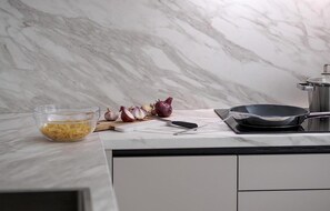 Eine Küchenarbeitsplatte mit einer Nudelschüssel, Knoblauch, Zwiebeln, einer Bratpfanne und einem Topf vor einer Marmorwand.
