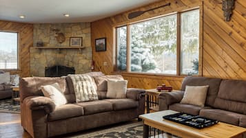 A cozy living room with two brown leather sofas, a stone fireplace, and a large window with a snowy view.