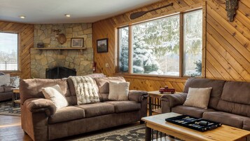 A cozy living room with two brown leather sofas, a stone fireplace, and a large window with a snowy view.