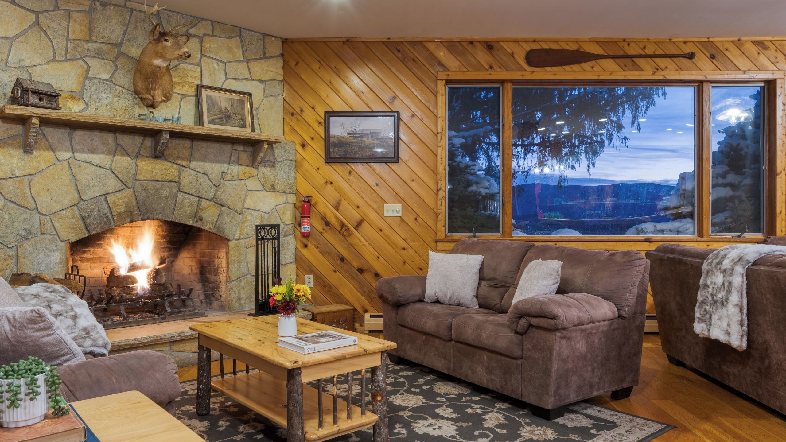 A cozy living room with a stone fireplace, wooden walls, and large windows offering a view of trees.