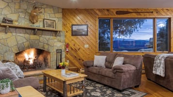 A cozy living room with a stone fireplace, wooden walls, and large windows offering a view of trees.