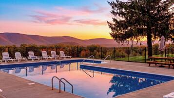 A swimming pool with lounge chairs and a picnic table, surrounded by a fence and trees, with a view of mountains in the background.