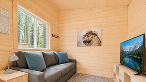 En mysig stuga med en soffa, en tv, ett skrivbord och ett fönster med utsikt över en skog.