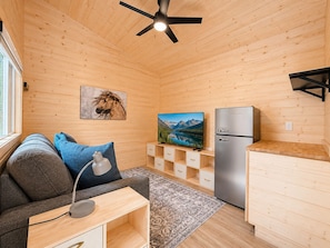 A cozy cabin interior with a wooden ceiling, a mounted TV, a refrigerator, a sofa with cushions, and a small table with a lamp.