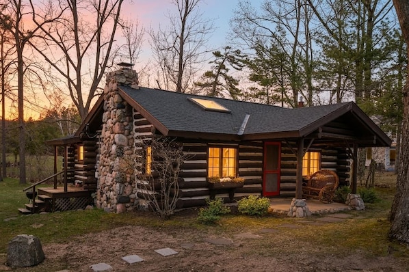 A log cabin with a stone chimney, a porch with a chair, and a window with warm lighting.