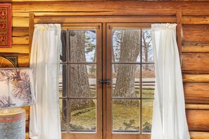 A wooden cabin with a double door window, white curtains, and a view of trees. - Dancing Bear Cabin: Authentic storybook log cabin with Lake Michigan views (Harbor Springs)