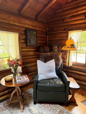 A cozy cabin interior with a leather chair, a small table with a candle, a vase of flowers, and a lamp with a decorative shade. - Dancing Bear Cabin: Authentic storybook log cabin with Lake Michigan views (Harbor Springs)