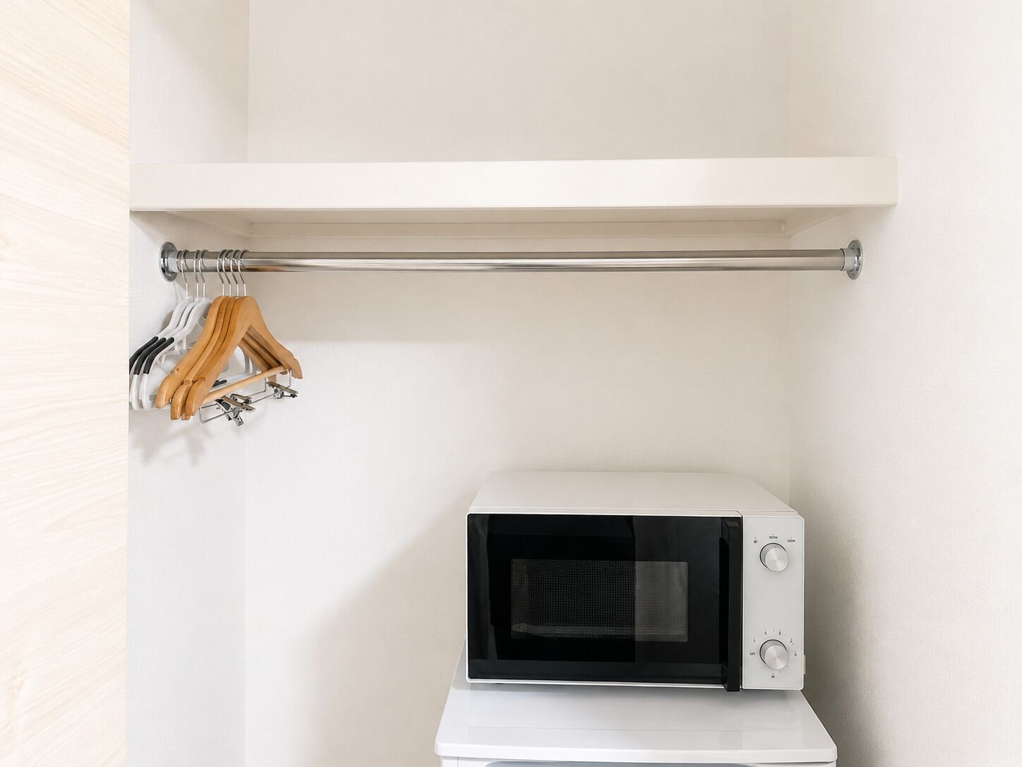 A white microwave oven on a white countertop, a white shelf above it, and a metal rod with wooden hangers.