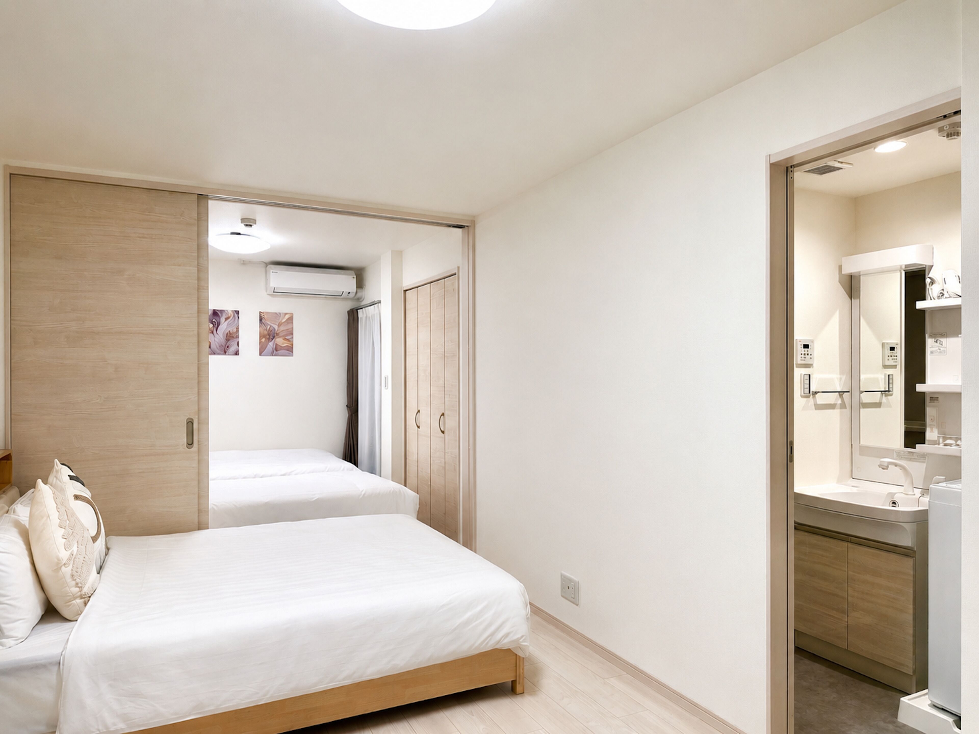 A bedroom with two beds, a wooden wardrobe, a bathroom with a mirror and sink, and an air conditioning unit.