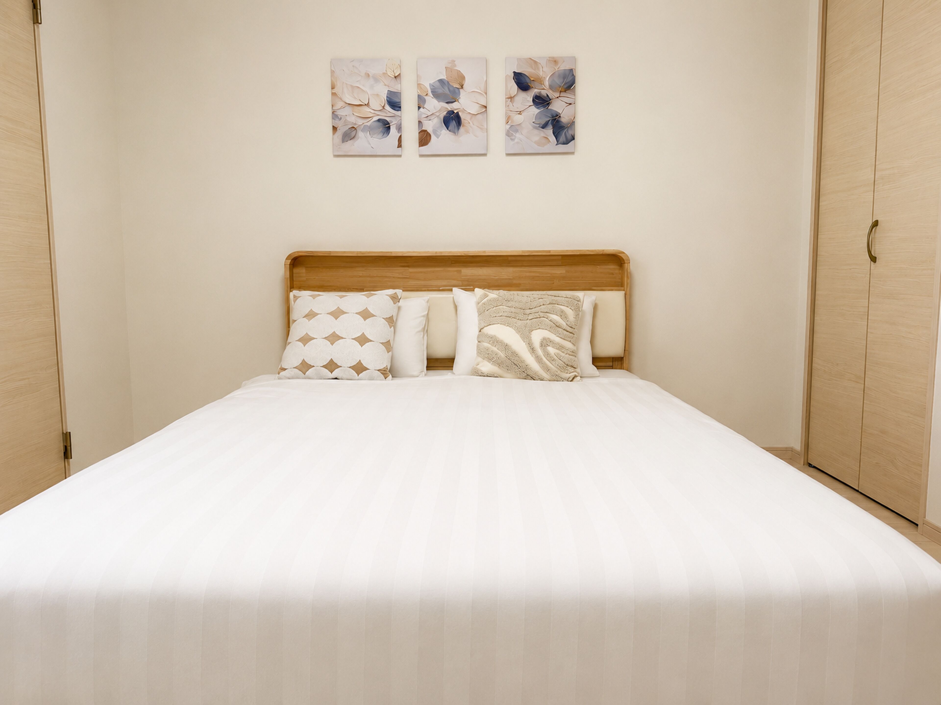 A neatly made bed with a wooden headboard, a plain wall with three abstract paintings, and a wooden wardrobe on the right.