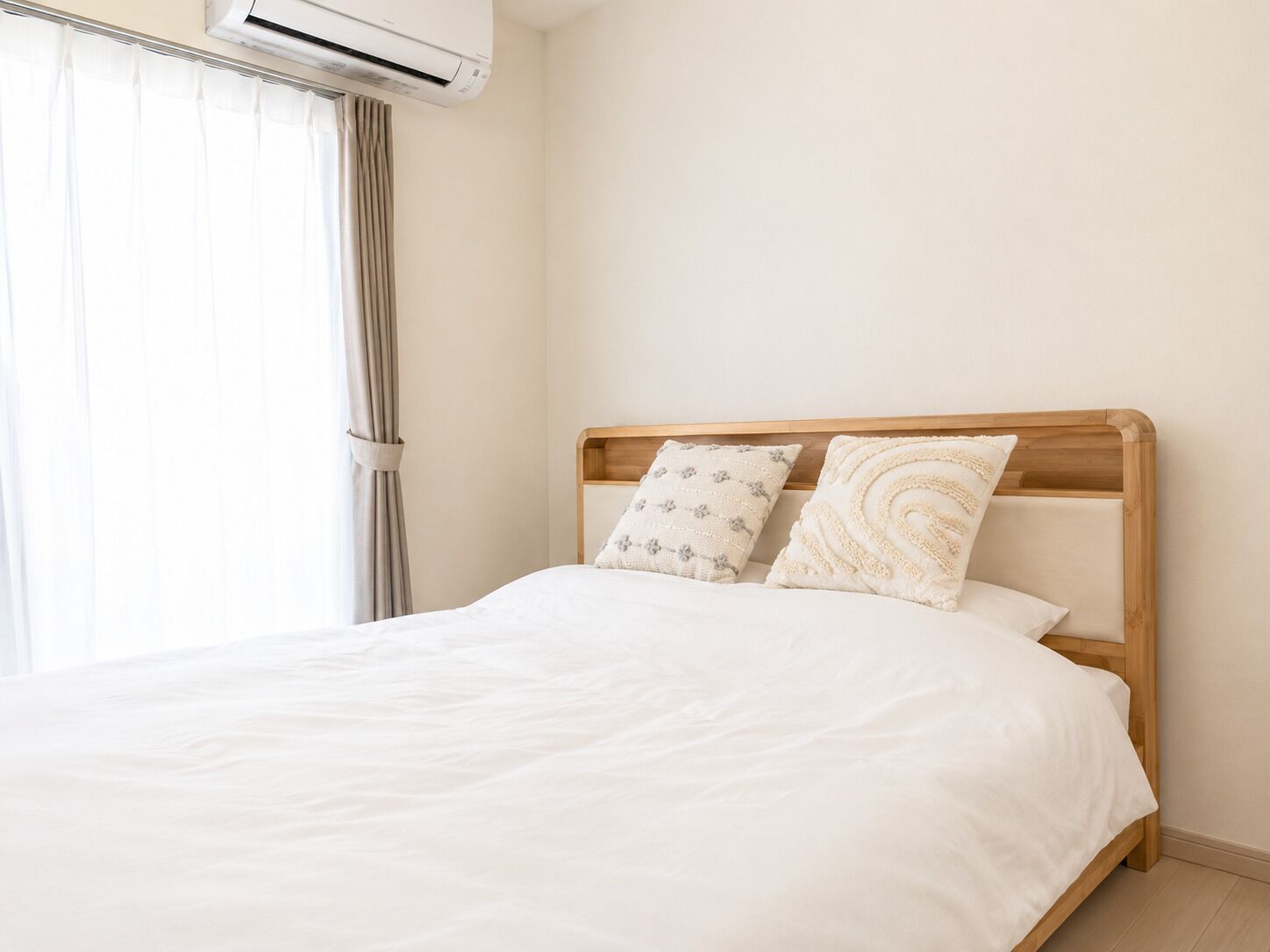 A neatly made bed with a wooden headboard, two patterned pillows, and a white bedspread in a room with a plain wall and an air conditioning unit above the window.