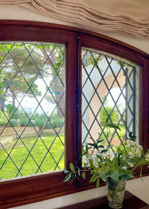 A window with a wooden frame and diamond-patterned glass, a potted plant on the windowsill, and a view of a grassy yard with trees. - Mediterranean villa with sea views, Lloret de Mar (Lloret de Mar)