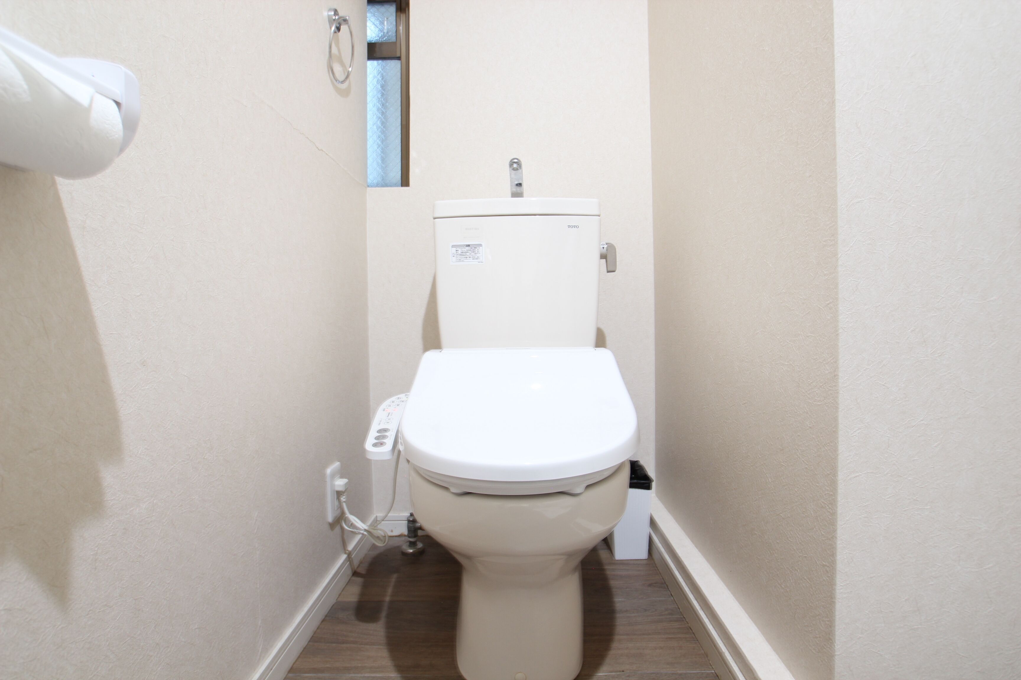 Een kleine, smalle badkamer met een wit toilet, een toiletrolhouder aan de muur en een zichtbaar stopcontact.