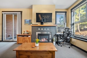 A modern living room with a fireplace, a dining table, a TV mounted above the fireplace, and a large window offering a view of trees.