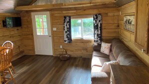 Innenansicht einer Holzhütte mit Holzboden, einer Couch, einem Esstisch, einem Stuhl, einem Fernseher und einem Fenster mit Vorhängen.
