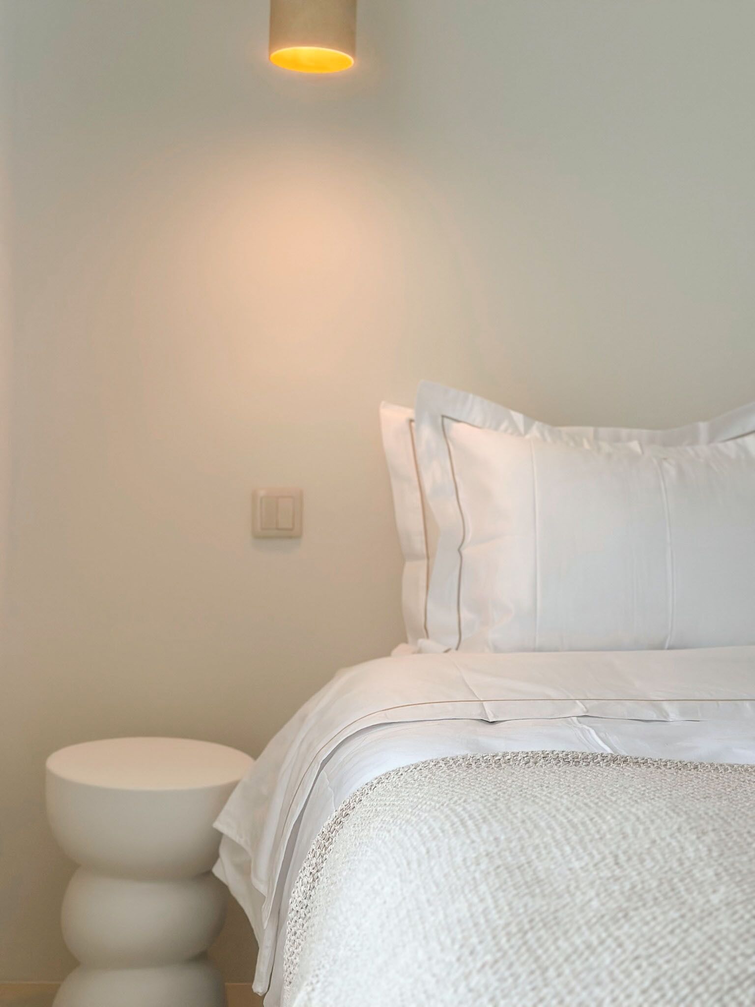 A bedroom with a bed, white pillows, a bedside table, and a wall-mounted light fixture.