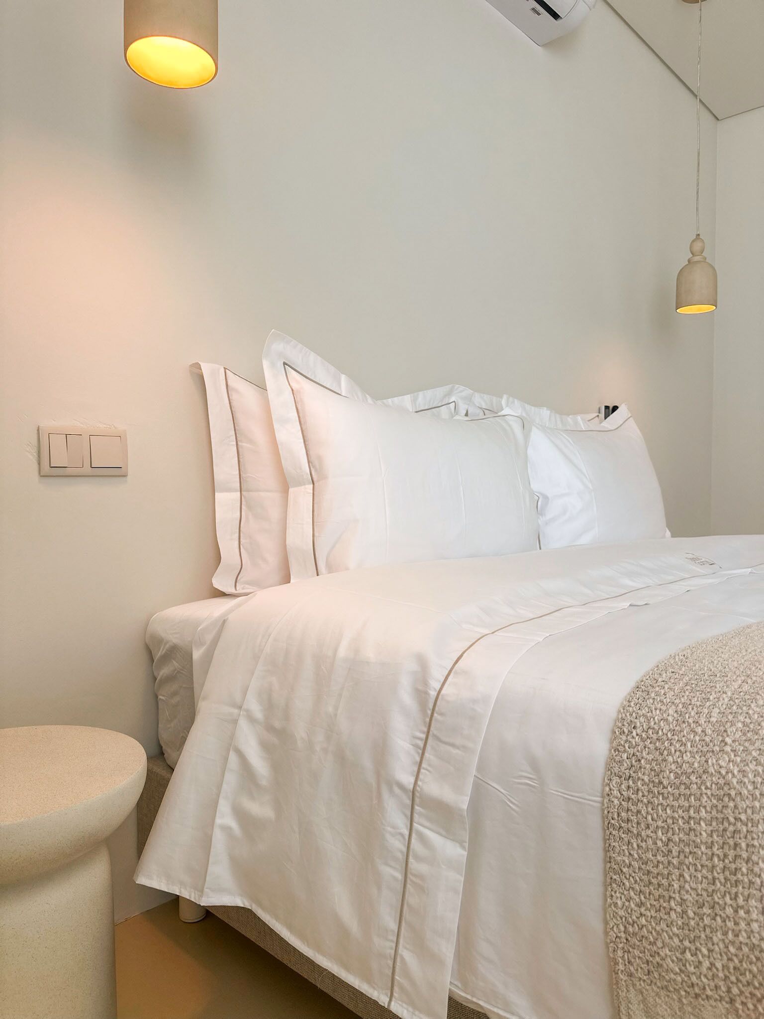 Cama bem arrumada com roupa de cama branca, luminária de cabeceira e ar condicionado fixado na parede.