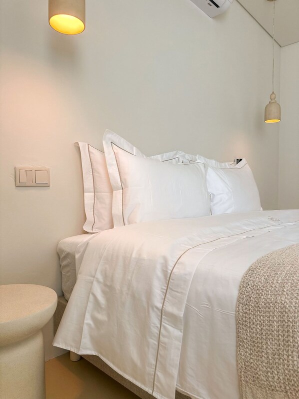 Una cama bien hecha con ropa blanca, una lámpara de noche y un aire acondicionado de pared.