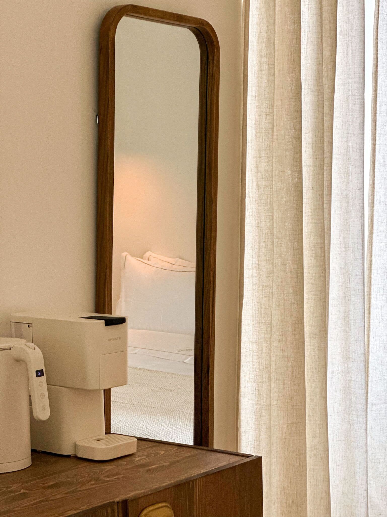 Et hotelværelse med et trænatbord, en hvid natlampe og et spejl der viser en seng med hvide sengetæpper.