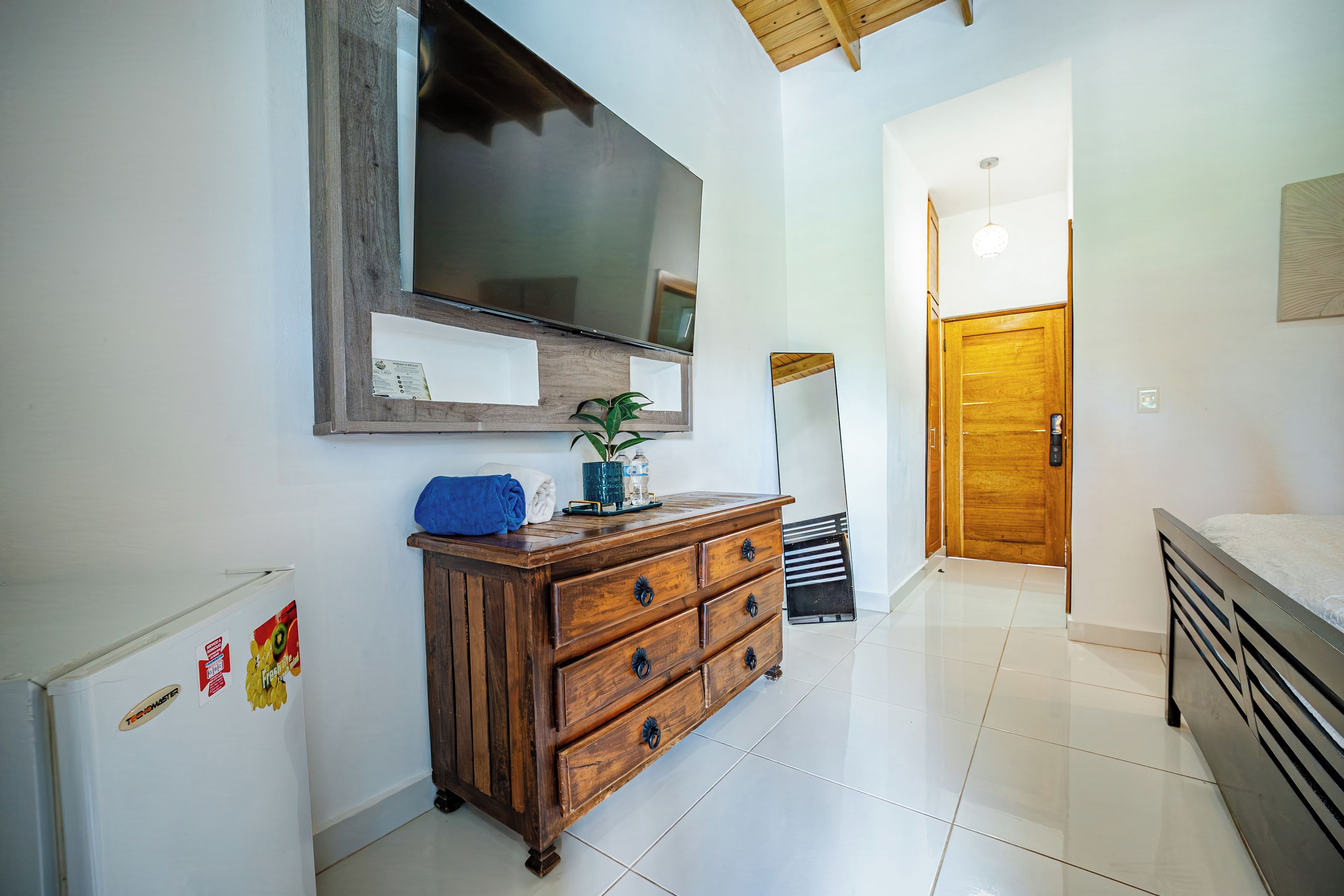 A bedroom with a wooden dresser, a bed, a large mirror, and a door leading to another room.