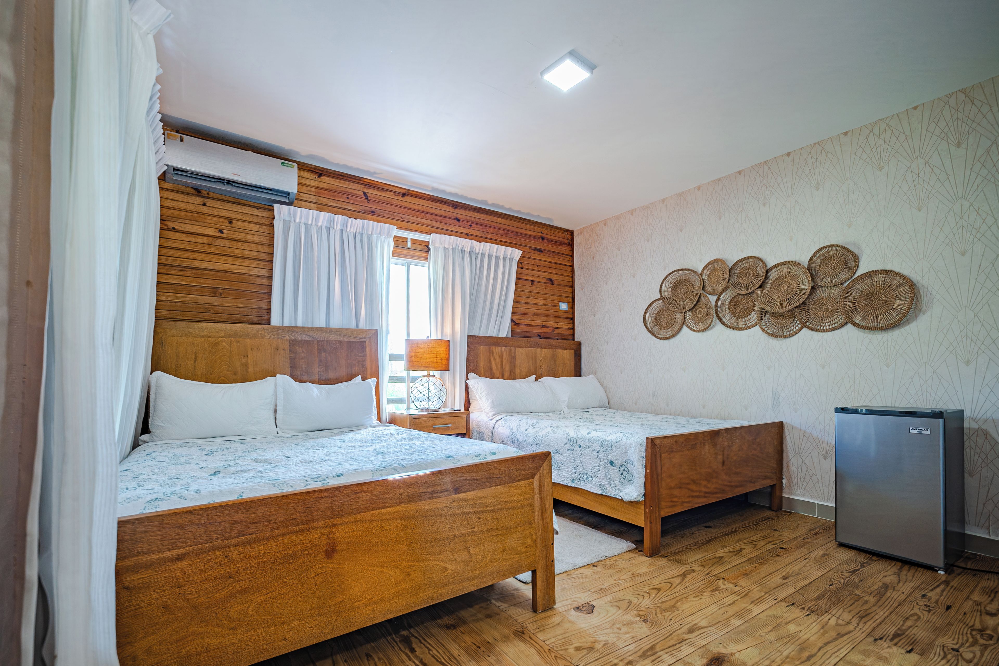 A bedroom with two wooden beds, a wall-mounted air conditioner, a refrigerator, and a decorative wall piece.