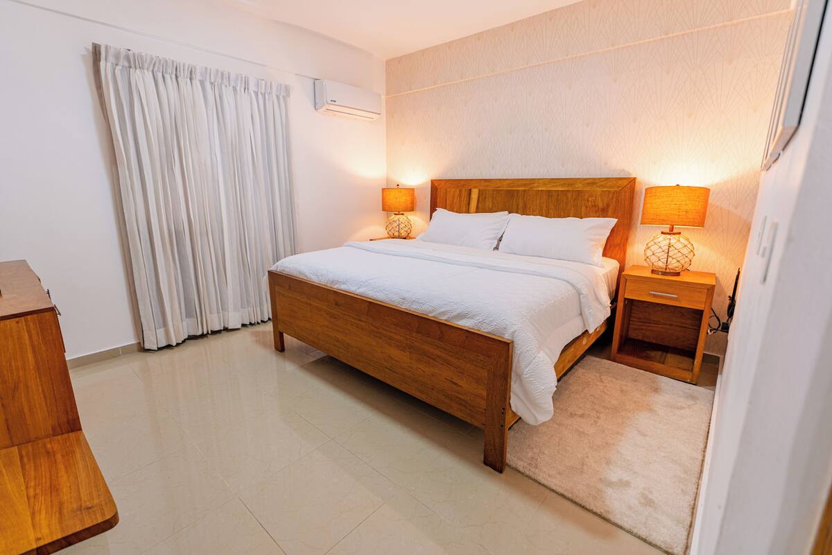 A neatly arranged bedroom with a wooden bed, two bedside tables with lamps, a window with curtains, and a wall-mounted air conditioner.