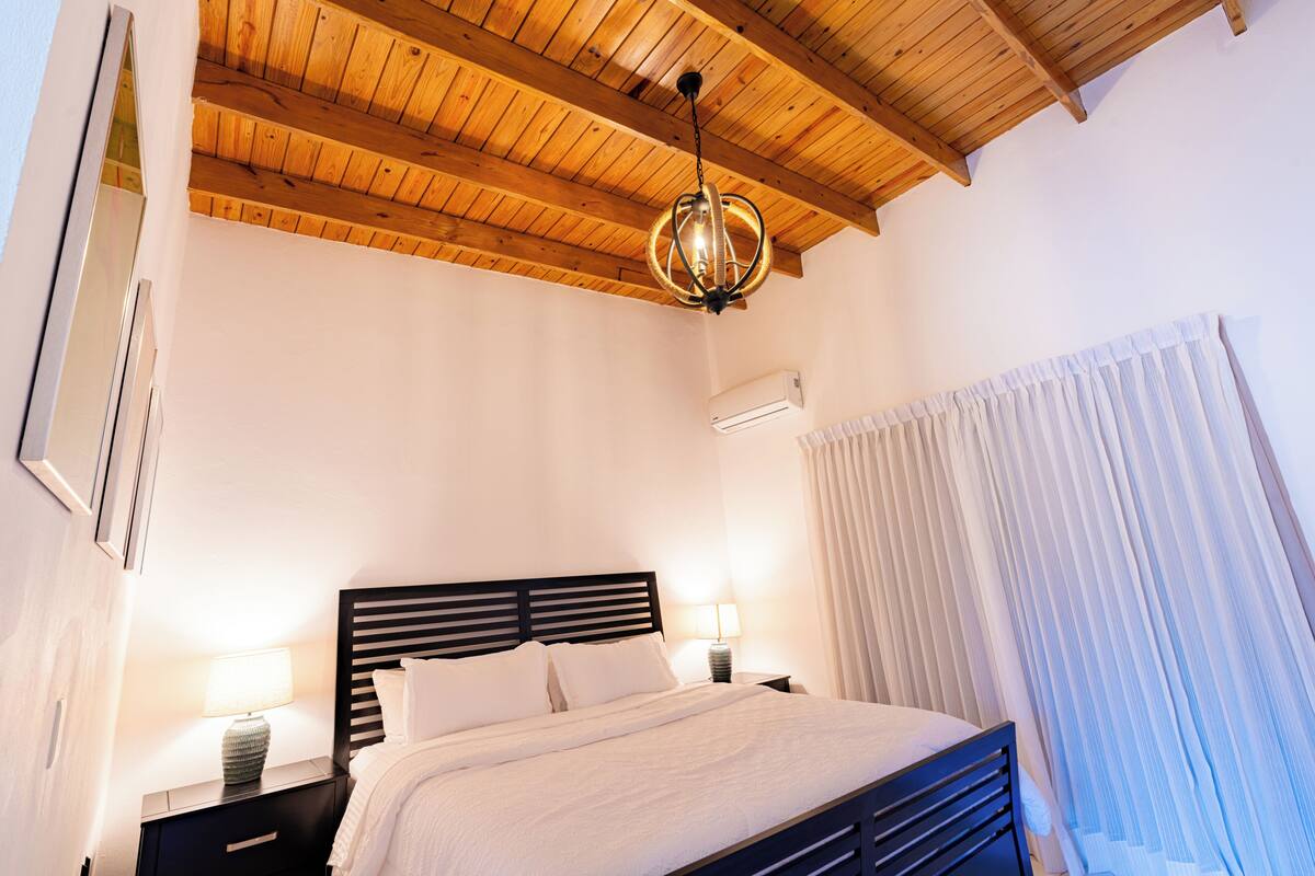 A bedroom with a wooden ceiling, a bed with white bedding, a nightstand with a lamp, and a window with curtains.
