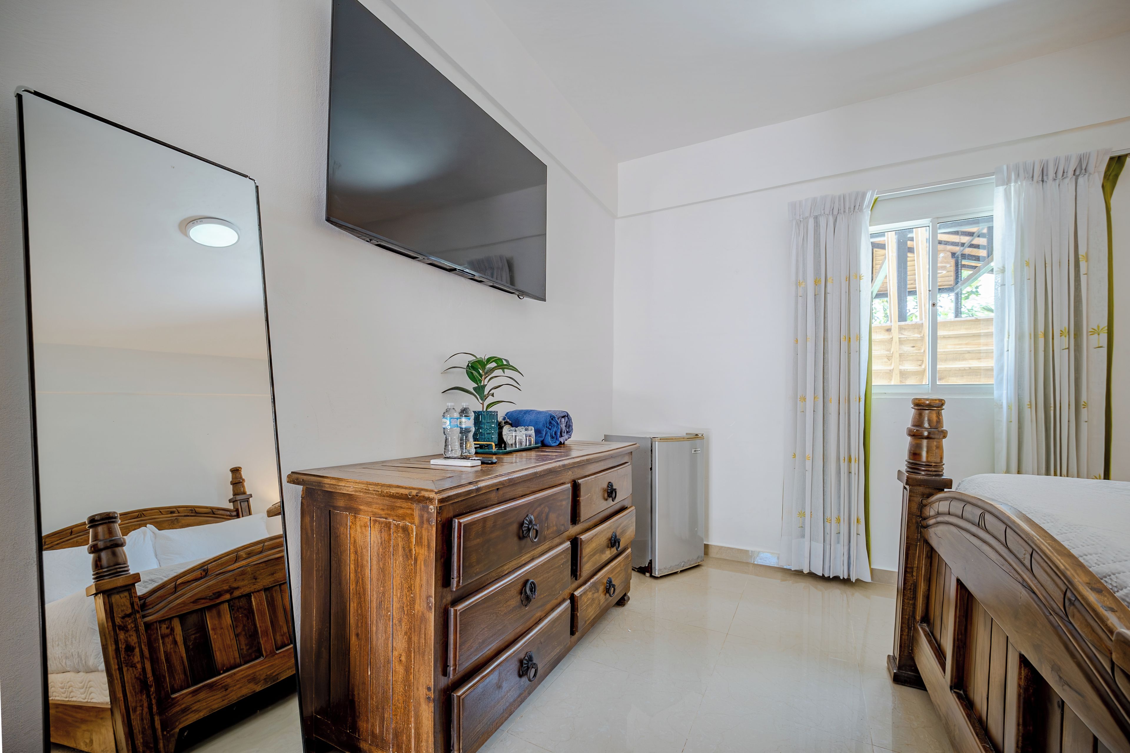Une chambre avec une commode en bois, un lit avec une tête de lit en bois, un téléviseur à écran plat fixé au mur, une fenêtre avec des rideaux et un radiateur.
