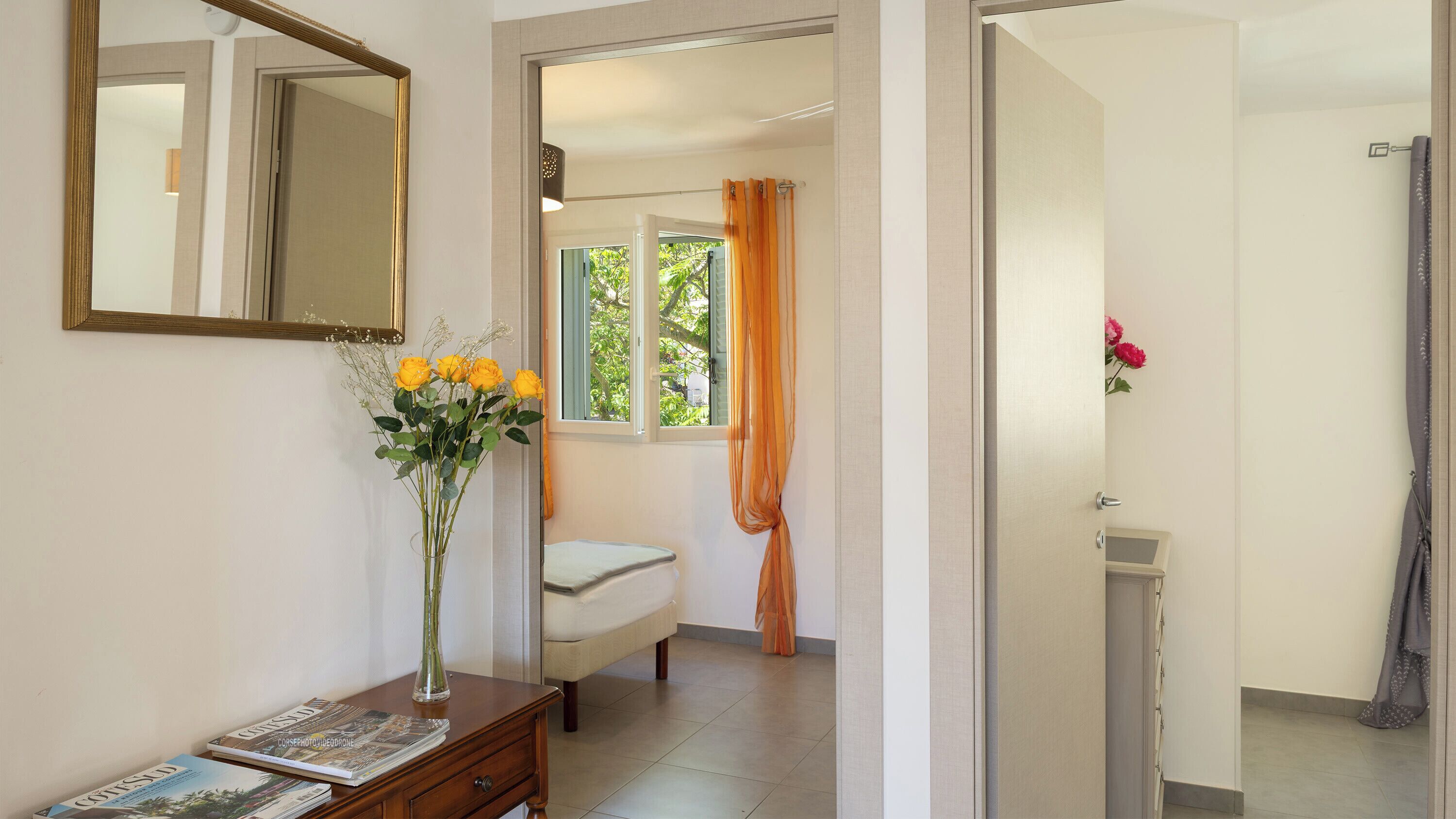 A room with a bed, a bedside table with a vase of flowers, a mirror, and a window with curtains.