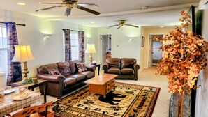 A living room with a brown leather sofa, a wooden coffee table, and a patterned rug. There are two ceiling fans, a lamp, and a decorative plant.