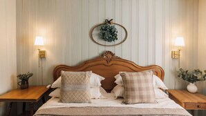 Una cama bien hecha con cabecera de madera, dos mesitas de noche con lámparas, una lámpara de pared y un arreglo decorativo en forma de corona.