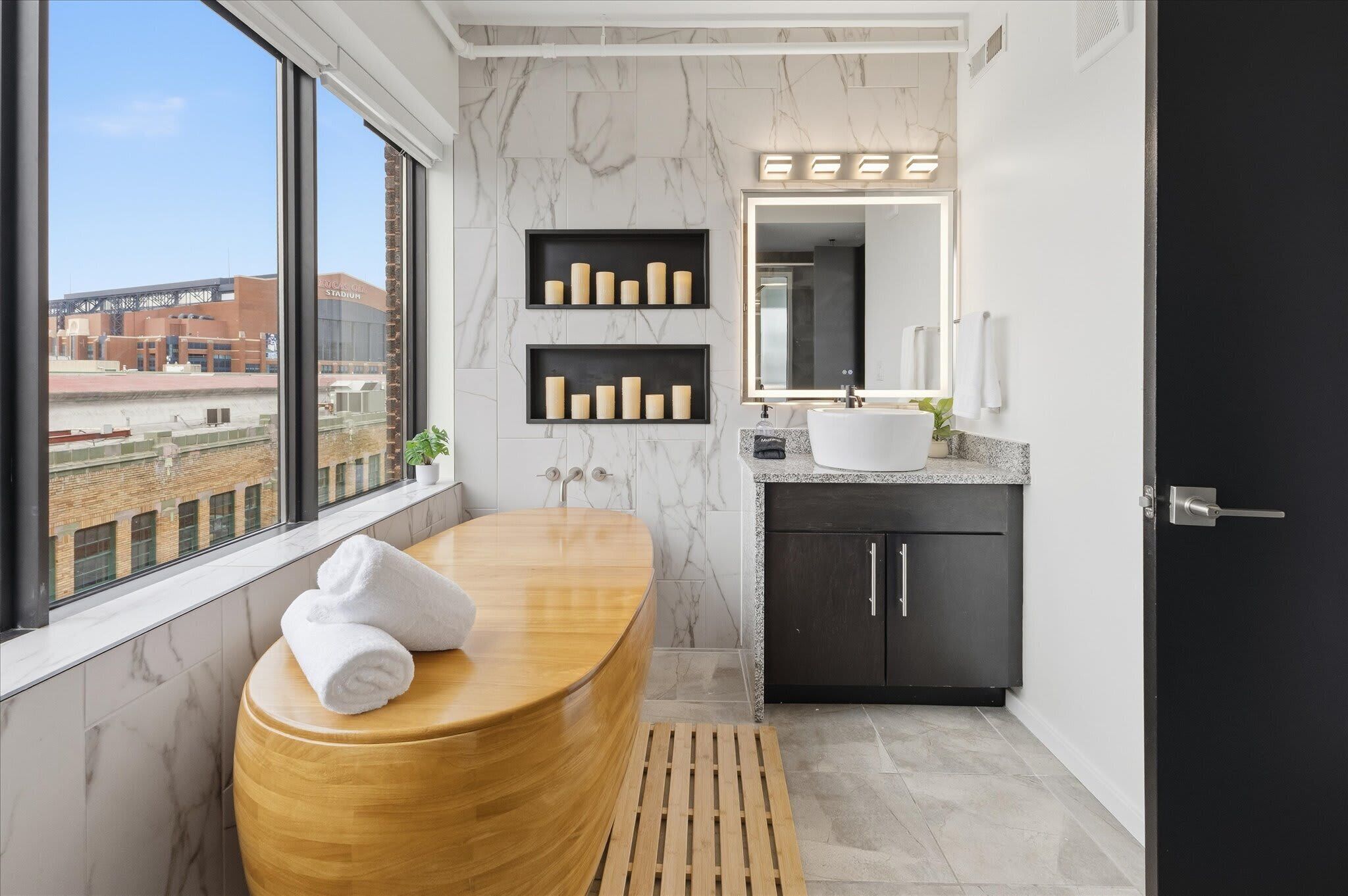 A modern bathroom with a wooden bathtub, marble walls, a large mirror, and a view of a stadium.