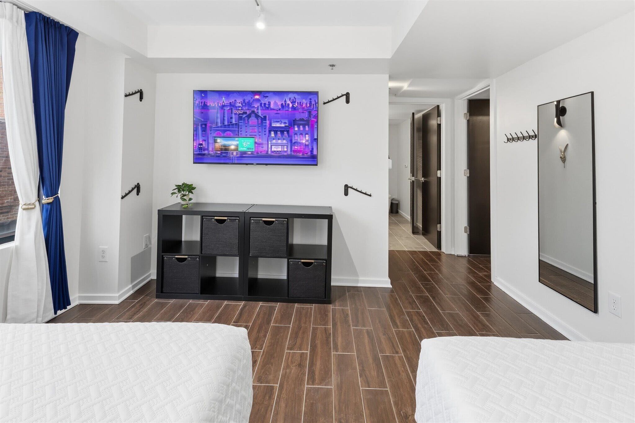 A hotel room with a TV mounted on the wall, a black storage unit, a bed, and a hallway leading to a door.