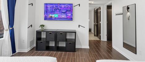 A hotel room with a TV mounted on the wall, a black storage unit, a bed, and a hallway leading to a door.