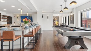 Una mesa de billar moderna con un soporte para tacos, una zona de bar con taburetes y un amplio ventanal con vistas a la ciudad.