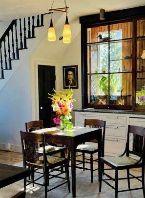 A dining area with a wooden table and chairs, a vase of flowers, a staircase with black balusters, and a window with plants.
