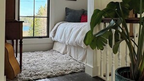A bedroom with a large window, a bed with white bedding, a guitar, a potted plant, and a framed picture on the wall.